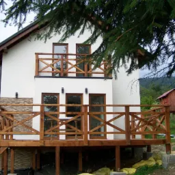 Dwukondygnacyjny budynek z białymi ścianami, drewnianymi balkonami i tarasem, w tle zalesione wzgórza i mały domek na palach.