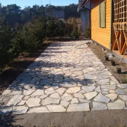 Ścieżka z nieregularnych, jasnych kamieni prowadząca do drewnianego domku z zielonymi okiennicami, obok posadzone młode świerki i doniczki z sadzonkami.