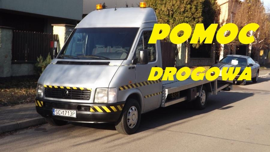 Srebrna laweta z żółto-czarnymi pasami holuje szary samochód osobowy na tle zabudowań mieszkalnych.