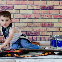 Chłopiec w bandanie i jeansach siedzi na podłodze z gitarą z motywem płomieni, obok bębenek zabawka, tło z cegieł