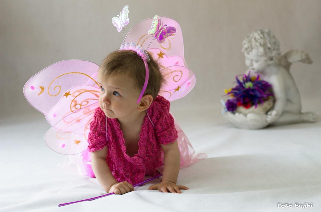 Mała dziewczynka w różowym stroju wróżki z motylkami na skrzydłach i różdżką, pozuje na białym tle z figurką anioła trzymającego kwiaty w tle.