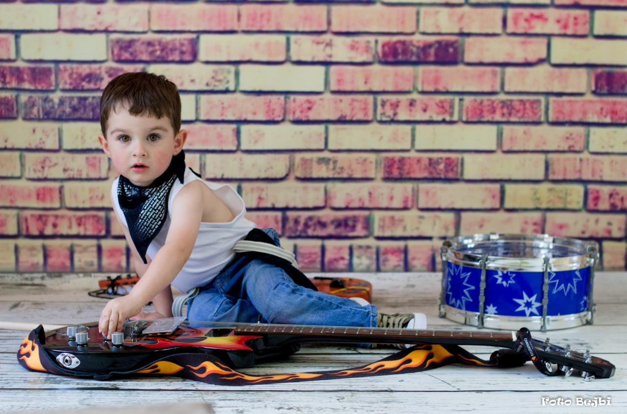 Chłopiec w bandanie i jeansach siedzi na podłodze z gitarą z motywem płomieni, obok bębenek zabawka, tło z cegieł