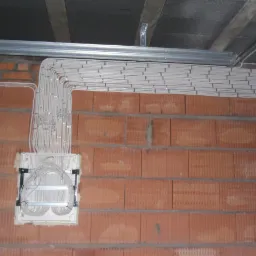 Równolegle ułożone kable elektryczne w białej osłonie prowadzone po czerwonej ścianie z cegły ceramicznej, w tle metalowa konstrukcja pod sufitem.