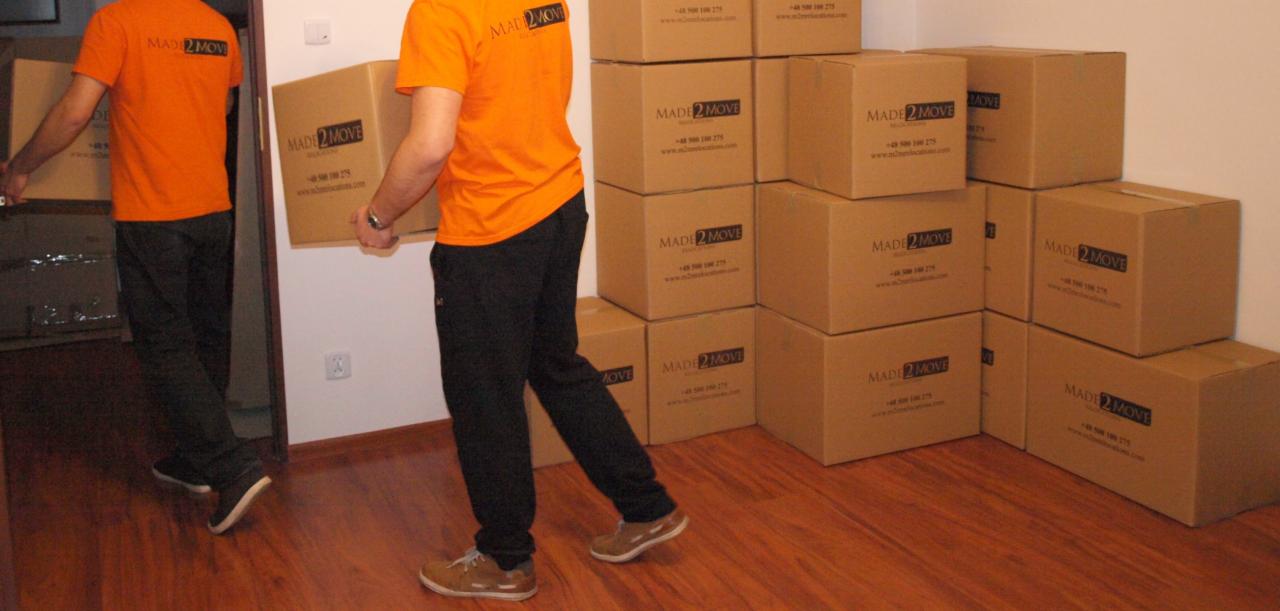 Pracownicy w pomarańczowych koszulkach z logo firmy transportowej niosą kartonowe pudła z logo firmy, obok ułożone stosy pudeł, widoczna podłoga z paneli.