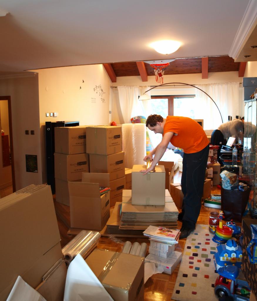 Mężczyzna w pomarańczowej koszulce zakleja karton taśmą w pokoju pełnym spakowanych pudeł, folii bąbelkowej i zabawek na dywanie w geometryczne wzory.