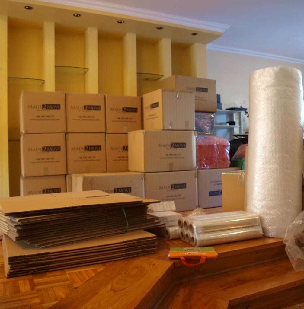 Stos kartonowych pudeł z nadrukiem 'MADE 2 MOVE', folia bąbelkowa i płaskie kartony przygotowane do pakowania na drewnianej podłodze w pomieszczeniu z żółtymi półkami.