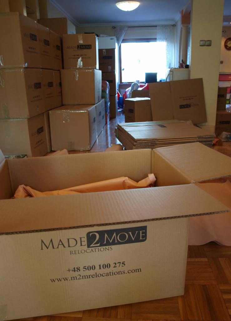 Karton z logo 'MADE 2 MOVE RELOCATIONS' wypełniony pomarańczowym materiałem, w tle sterta kartonów w pokoju przed oknem.