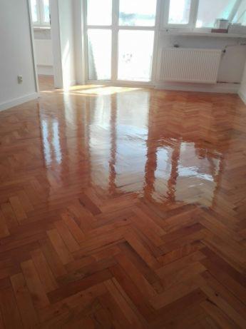 Błyszcząca, świeżo wykończona podłoga z parkietu ułożonego we wzór jodełki, odbijająca światło z okna w jasnym pomieszczeniu.