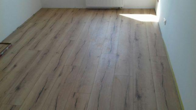 Nowo ułożone panele podłogowe w jasnym odcieniu drewna z widocznymi, ciemnymi słojami, częściowo przykryte kartonem, w pomieszczeniu z białymi ścianami i grzejnikiem.