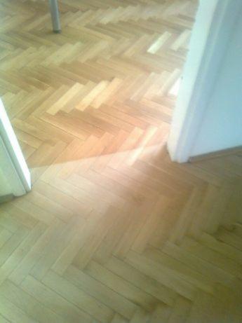 Jasny parkiet ułożony we wzór jodełki, widoczny fragment futryny drzwi i noga stołu. Świeżo położona podłoga w mieszkaniu.