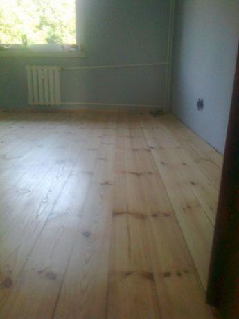Jasne, świeżo wycyklinowane deski podłogowe z widocznymi śladami po szlifowaniu, w tle grzejnik i szara ściana z widocznymi przewodami elektrycznymi.