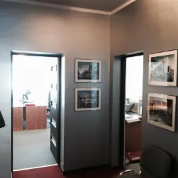 Korytarz biurowy w Gdyni z szarą ścianą, na której wiszą dwie pary oprawionych zdjęć. W tle widoczne biurka i krzesła w pomieszczeniach biurowych.