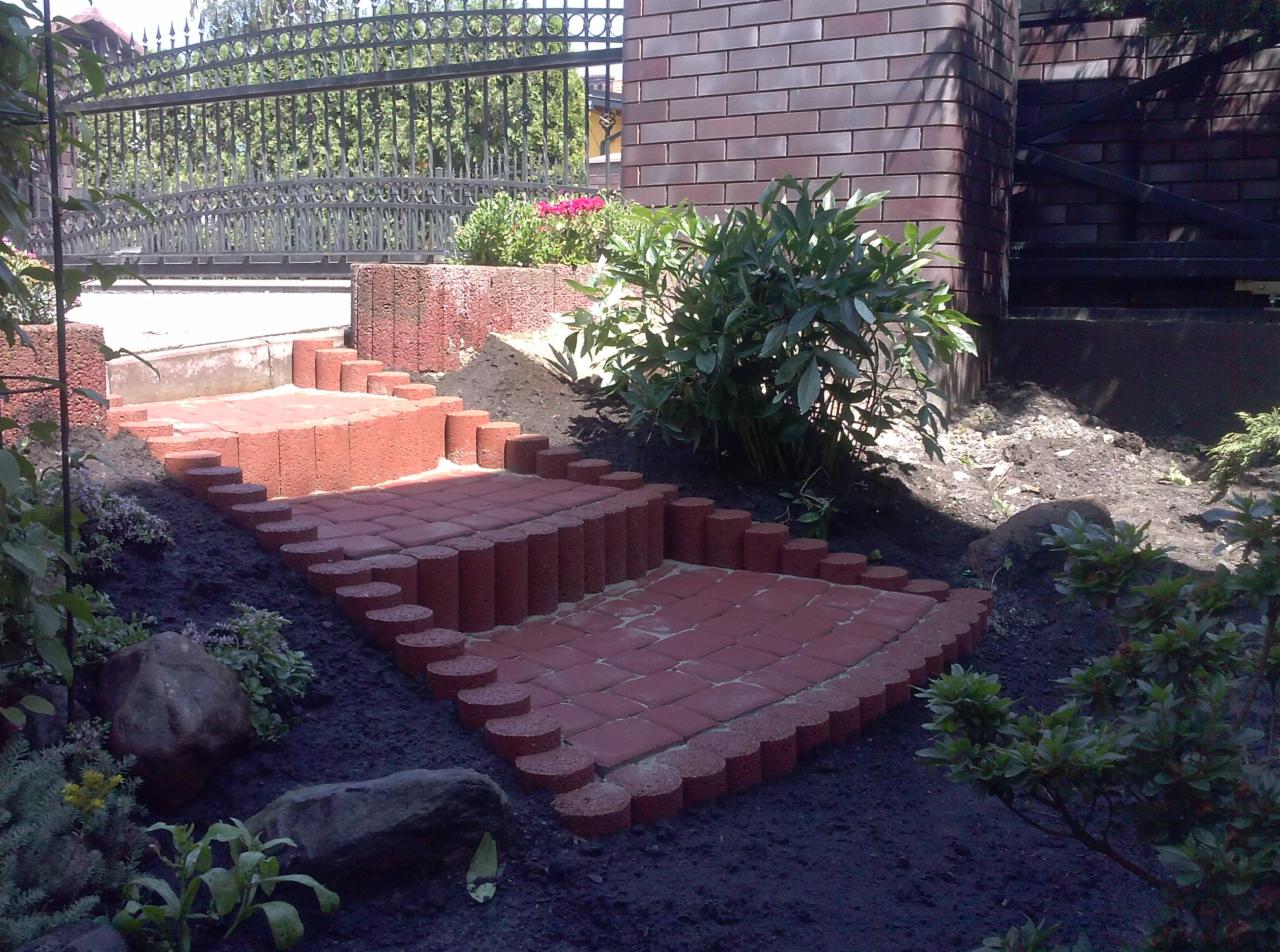 Schody ogrodowe z czerwonej kostki brukowej z obrzeżami z okrąglaków, prowadzące przez rabatę w kierunku bramy z kutego żelaza.