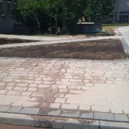 Nowo ułożona, jasnoszara kostka brukowa z ciemniejszym obramowaniem, tworząca ścieżkę i plac. Widoczne fragmenty rabat z ziemią przygotowanych pod nasadzenia. Część kostki jest jeszcze mokra po...