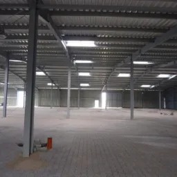 Wnętrze nowej hali z widoczną konstrukcją stalową, betonową posadzką z kostki i świetlikami dachowymi, widoczne przyłącza instalacyjne przy słupach.