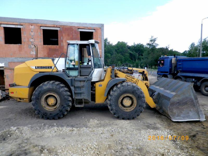 Żółta ładowarka kołowa Liebherr 554 na placu budowy, z widocznym budynkiem w trakcie wznoszenia z cegły dziurawki w tle oraz niebieską ciężarówką wywrotką po prawej stronie.