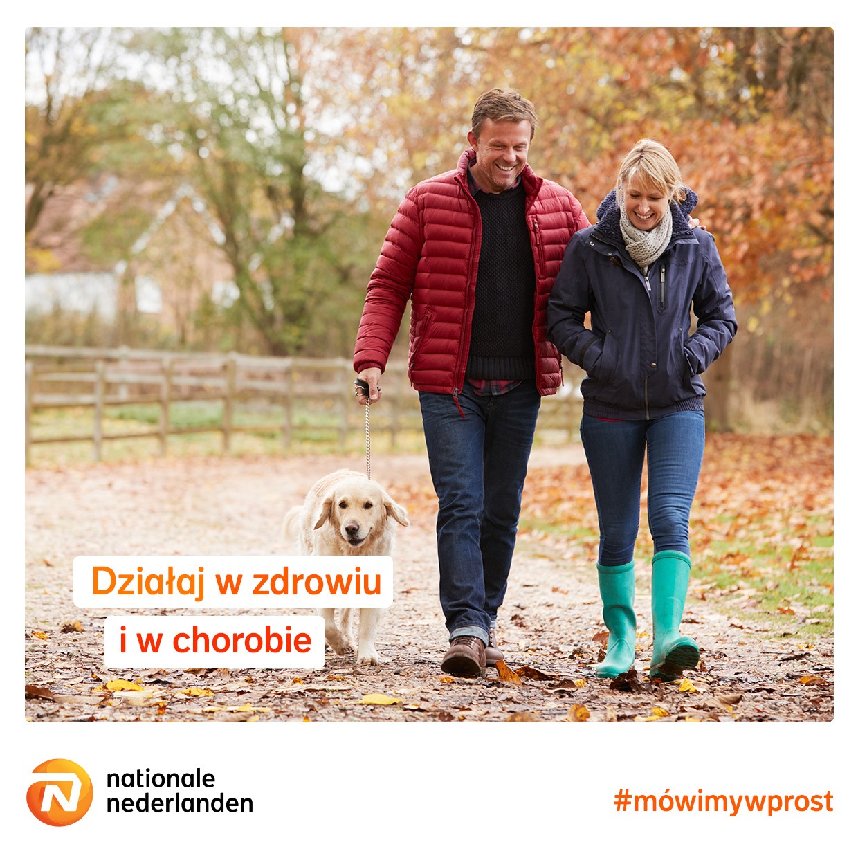 Uśmiechnięta para spaceruje jesienną aleją z psem na smyczy. Kobieta w turkusowych kaloszach, mężczyzna w czerwonej kurtce.