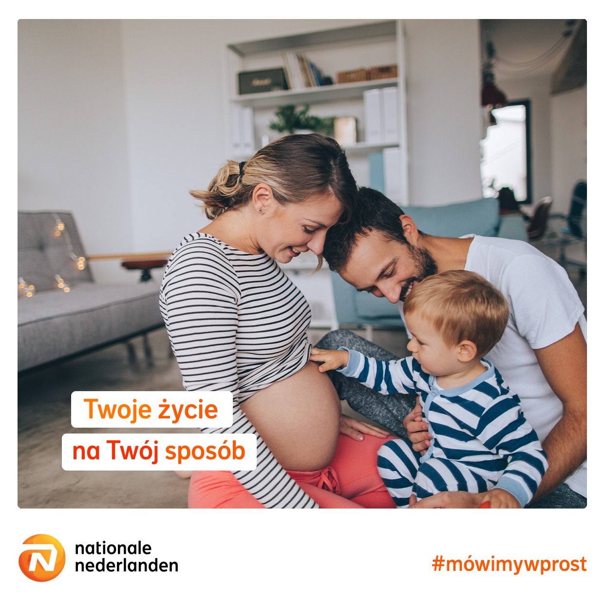 Uśmiechnięta, ciężarna kobieta w bluzce w paski, ojciec i mały syn dotykający brzucha, w tle jasny salon z regałem i kanapą.