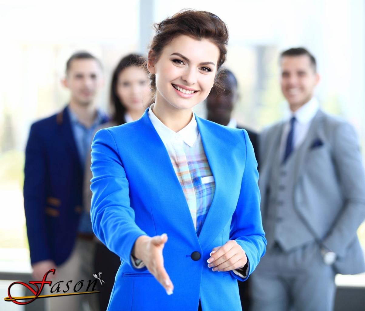 Uśmiechnięta kobieta w niebieskim żakiecie wyciąga rękę na powitanie, w tle grupa osób w strojach biznesowych i logo firmy odzieżowej w lewym dolnym rogu.