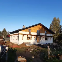 Dom jednorodzinny po częściowym wykończeniu elewacji, z drewnianą górną częścią i białą dolną, widoczne rusztowania i materiały budowlane na pierwszym planie.