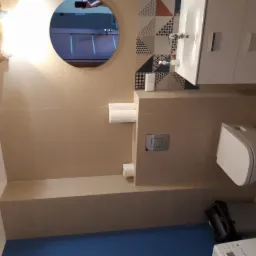 Beżowa łazienka z geometryczną mozaiką nad umywalką i okrągłym lustrem, w którym odbija się fragment innego pomieszczenia.