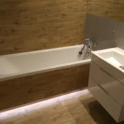 Nowoczesna łazienka z wanną obudowaną płytkami imitującymi drewno, białą umywalką z szafką i szarą ścianą, oświetlona listwą LED pod wanną.