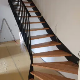 Nowoczesne schody z drewnianymi stopniami i czarną, metalową balustradą, widoczne zabezpieczenie podłogi folią malarską i drabina w tle, sugerujące trwający remont.