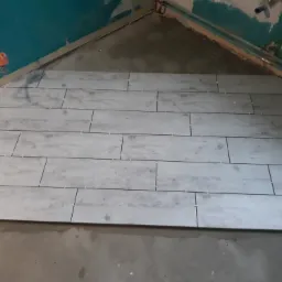 Ułożone płytki podłogowe imitujące drewno w rogu pomieszczenia, z widocznymi krzyżykami dystansowymi i fragmentem nieukończonej podłogi.