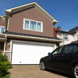 Nowoczesny dom jednorodzinny z brązową elewacją i białą, segmentową bramą garażową, obok zaparkowany czarny samochód osobowy.