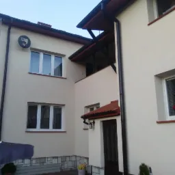 Dwupiętrowy dom z jasną, gładką elewacją, widoczne okna z białymi ramami, czarne rynny, balkon z drewnianą konstrukcją, dolna część ściany wykończona kamieniem dekoracyjnym, satelita na ścianie.