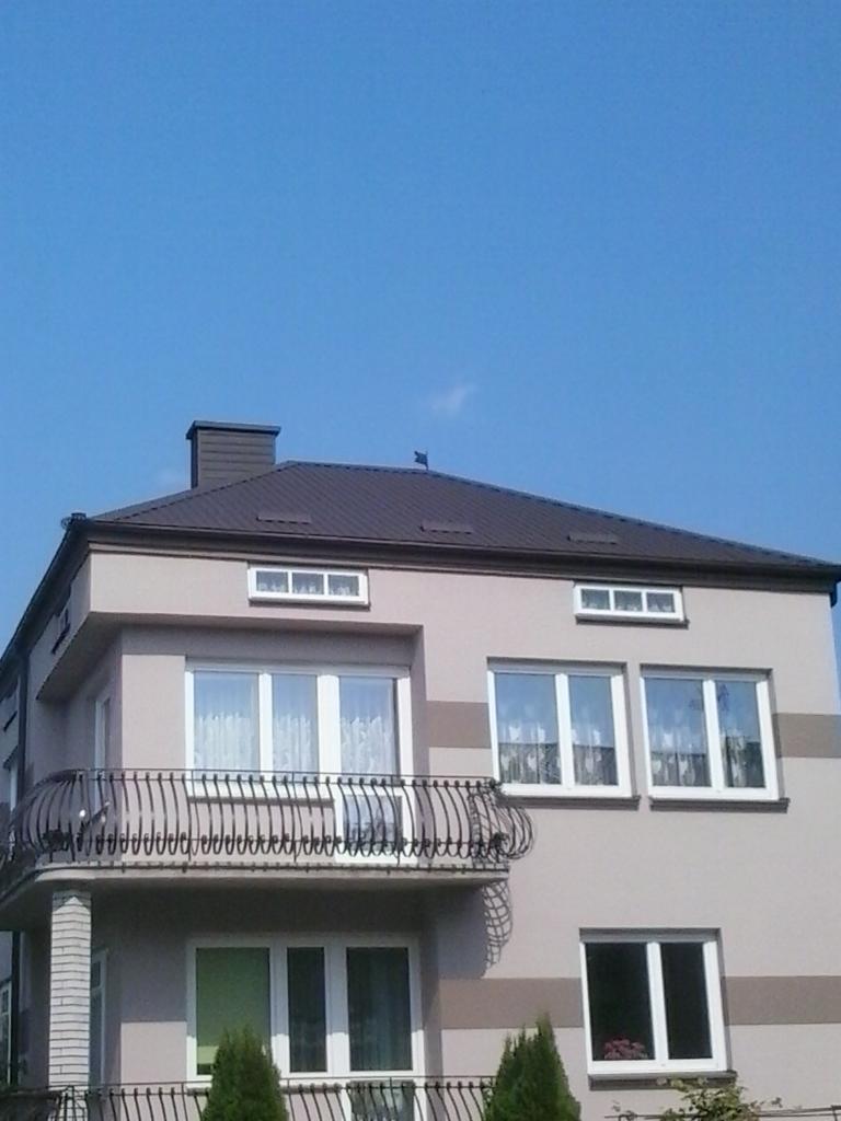 Dwupiętrowy dom z brązowym dachem i balkonem z kutą balustradą, szara elewacja z poziomymi pasami, widok z dołu na tle błękitnego nieba.