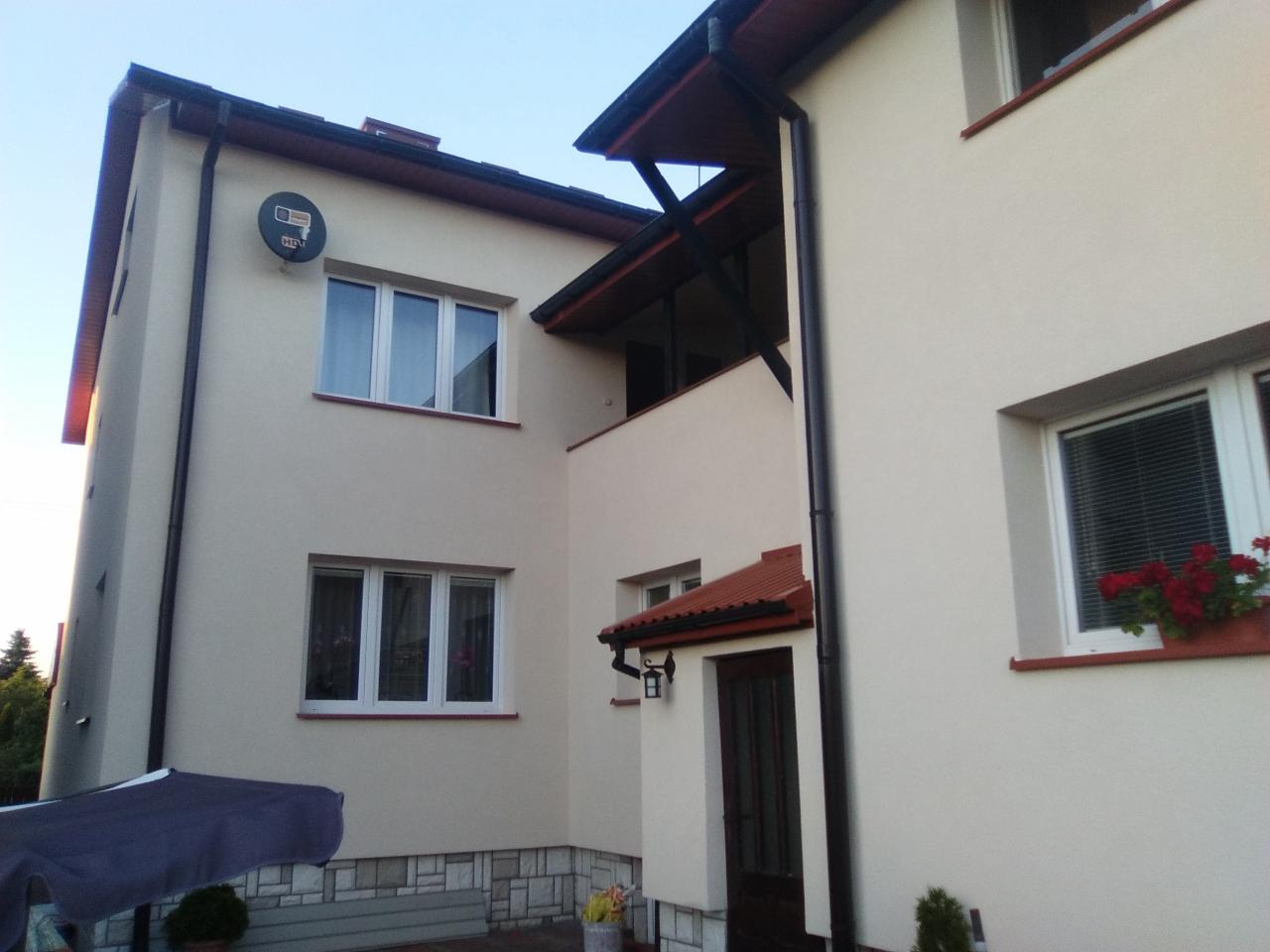 Dwupiętrowy dom z jasną, gładką elewacją, widoczne okna z białymi ramami, czarne rynny, balkon z drewnianą konstrukcją, dolna część ściany wykończona kamieniem dekoracyjnym, satelita na ścianie.
