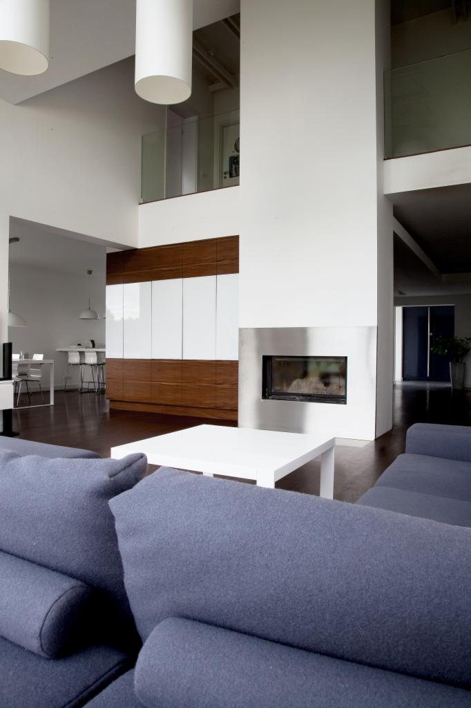 Wnętrze nowoczesnego domu z antresolą, widok na salon z szarej sofy, biały stolik kawowy, kominek w metalowej obudowie oraz zabudowę meblową z drewnianymi i białymi frontami.