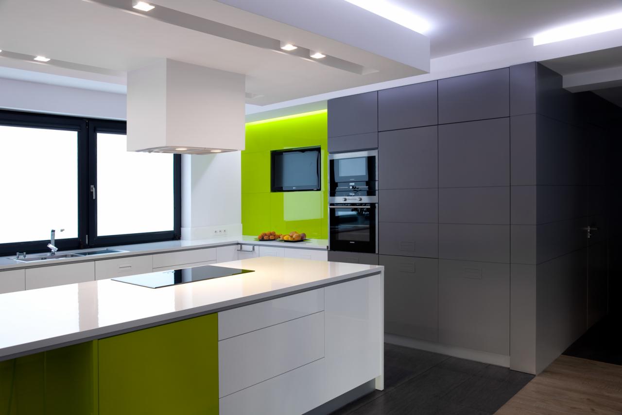 Nowoczesna kuchnia z białymi i zielonymi szafkami, wyspą z płytą indukcyjną, szarymi szafkami w zabudowie, zielonym panelem szklanym na ścianie i wiszącym telewizorem.
