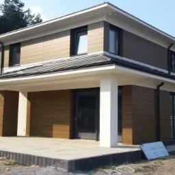 Dwukondygnacyjny dom z brązową elewacją imitującą drewno, białymi elementami wykończeniowymi i szarym dachem, otoczony zielenią i częściowo wykończonym terenem.