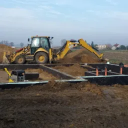 Przygotowanie fundamentów pod budynek: wykopane i zabezpieczone wykopy, widoczny sprzęt budowlany i robotnik.