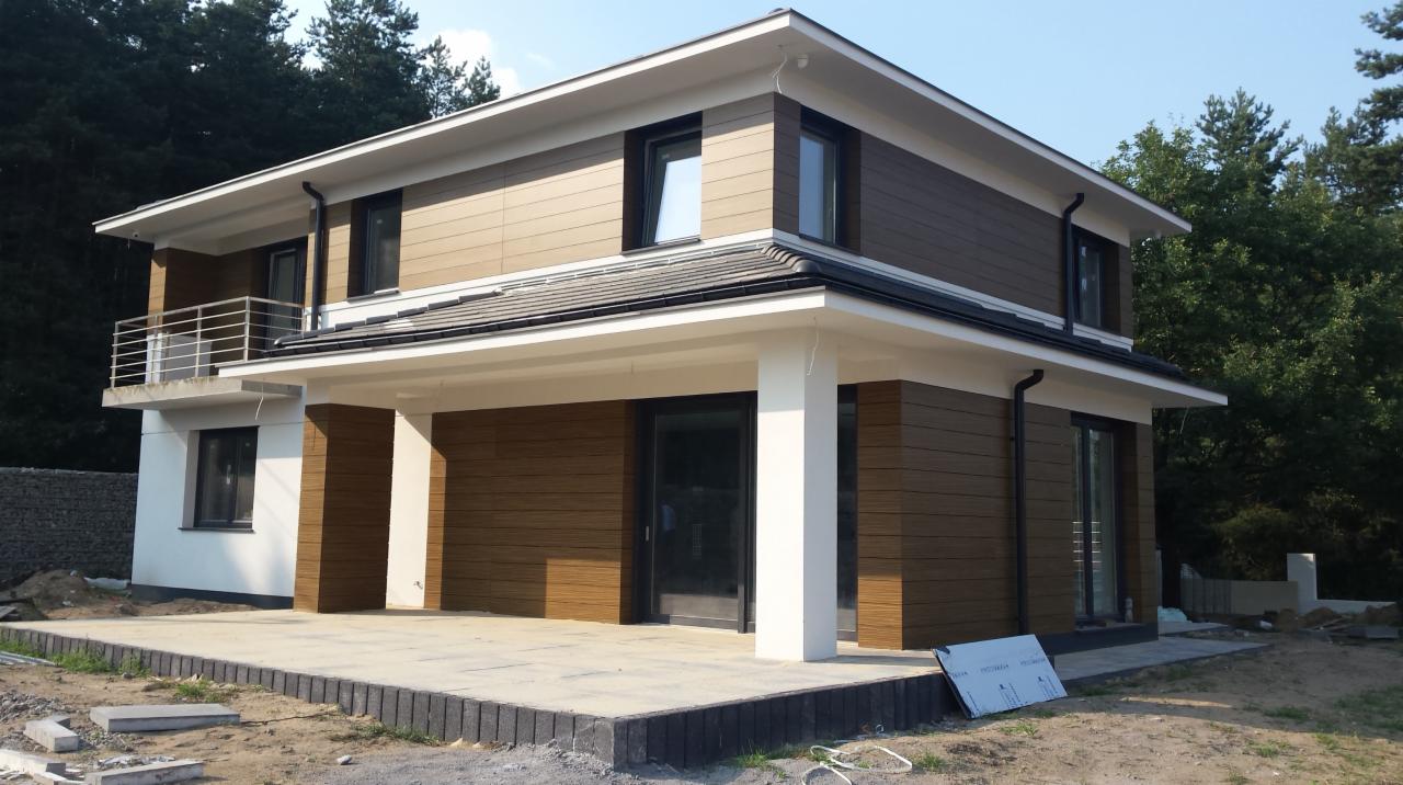 Dwukondygnacyjny dom z brązową elewacją imitującą drewno, białymi elementami wykończeniowymi i szarym dachem, otoczony zielenią i częściowo wykończonym terenem.