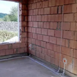 Wnętrze budynku w trakcie budowy z widoczną instalacją elektryczną: kable w listwach przypodłogowych oraz puszka elektryczna w ścianie z czerwonej cegły dziurawki.