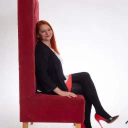 Kobieta z rudymi włosami siedzi na bardzo wysokim, czerwonym fotelu w czarnej marynarce i czarnych rajstopach, pozuje na białym tle, ma na sobie czarne szpilki z czerwonym obcasem.