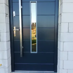Nowoczesne drzwi zewnętrzne w kolorze grafitowym z pionowym przeszkleniem i długim, srebrnym uchwytem, zamontowane w murze z białych bloczków.