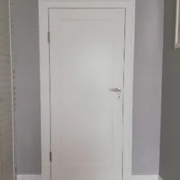 Białe drzwi z delikatnym frezem i srebrną klamką na tle szarej ściany i podłogi imitującej drewno.