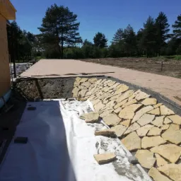 Budowa tarasu z kamienną ścianką oporową i wyłożoną kostką ścieżką, widok z góry na zabezpieczoną folią przestrzeń przy budynku.