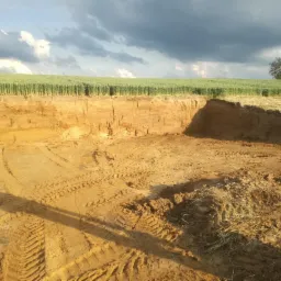 Rozległy wykop w ziemi z widocznymi śladami opon maszyn budowlanych, w tle pole uprawne i pochmurne niebo.