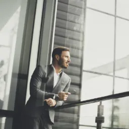 Uśmiechnięty mężczyzna w szarym garniturze, oparty o balustradę, trzyma telefon i spogląda przez okno w nowoczesnym biurowcu.