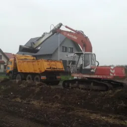 Czerwono-biała koparka RHS5 PMS ładuje ziemię do żółtej ciężarówki na tle budynków mieszkalnych w pochmurny dzień.