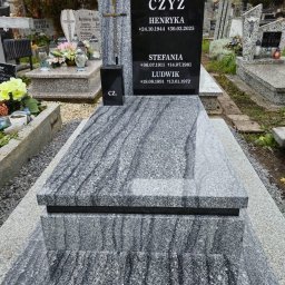 KAMIENIARSTWO Teresa Kwiatkowska - Granitowy nagrobek z czarną tablicą inskrypcyjną i krzyżem, otoczony żwirem na cmentarzu. Widoczne inne nagrobki w tle, jesienne drzewa.