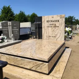 Jasny, beżowo-brązowy nagrobek z granitu z ciemnym krzyżem i napisem KUCHAREK ZOFIA, na cmentarzu, w tle inne nagrobki i drzewa.