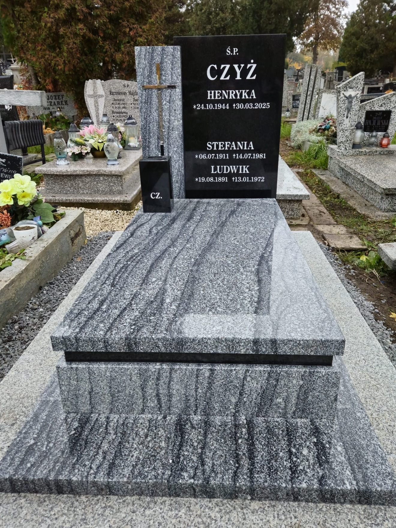Granitowy nagrobek z czarną tablicą inskrypcyjną i krzyżem, otoczony żwirem na cmentarzu. Widoczne inne nagrobki w tle, jesienne drzewa.