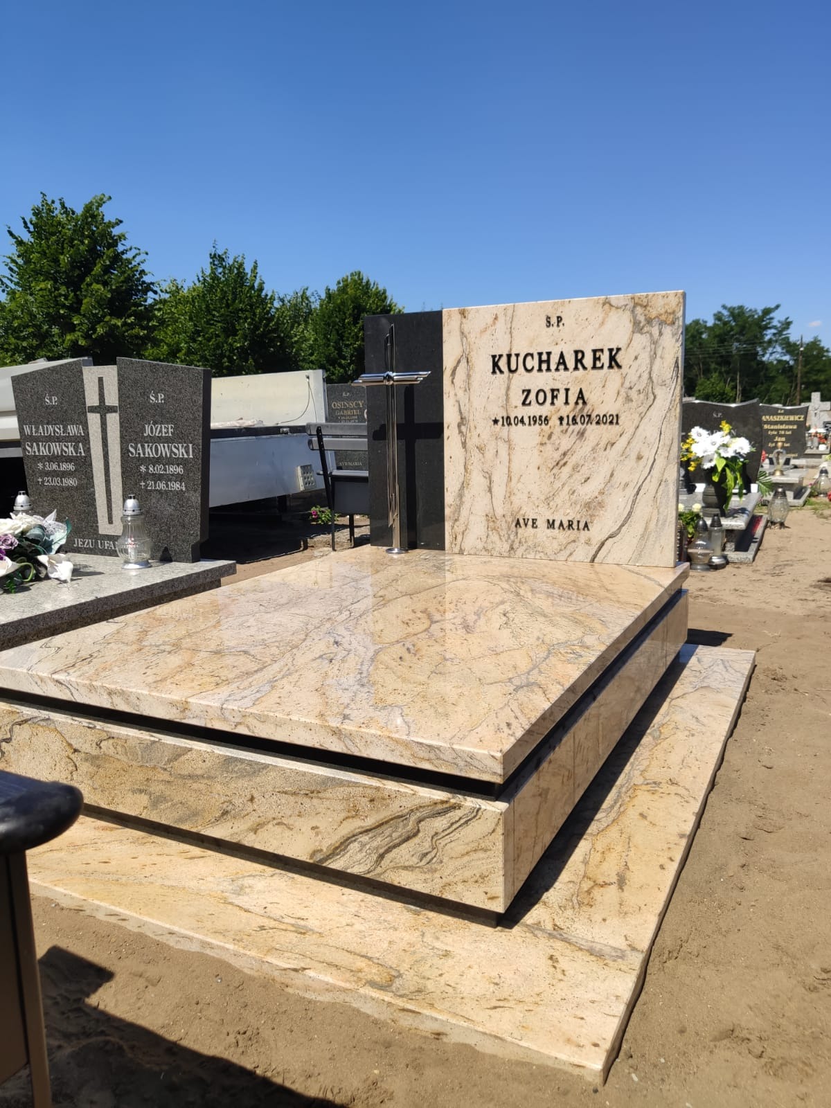 Jasny, beżowo-brązowy nagrobek z granitu z ciemnym krzyżem i napisem KUCHAREK ZOFIA, na cmentarzu, w tle inne nagrobki i drzewa.