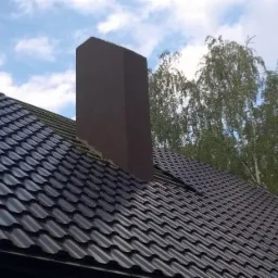 Nowy, ciemny dach z blachodachówki z brązowym kominem na tle błękitnego nieba i drzew